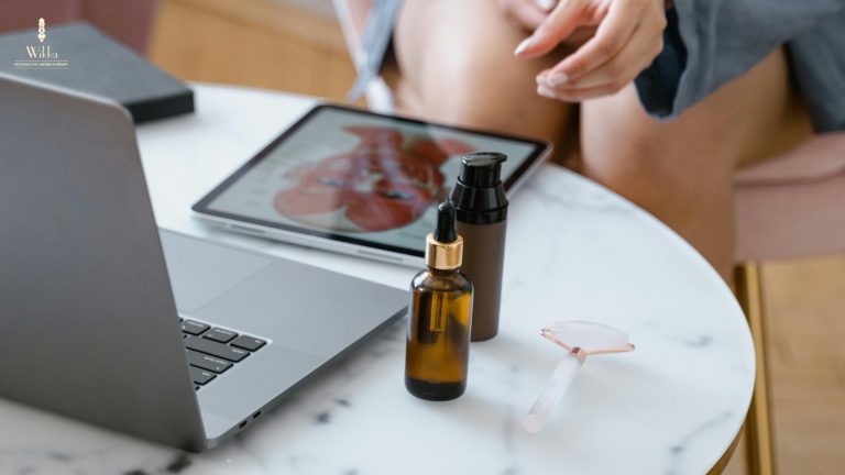 Aromatherapy essential oil bottles, rose quartz roller, and tablet on a marble desk beside a laptop, promoting digital wellness and self-care.